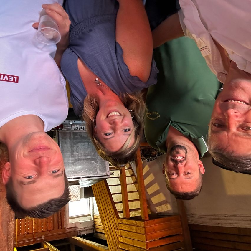 Drei Personen machen ein Selfie im Dachboden, lächeln und schauen in die Kamera. Ein Mann trägt ein weißes Hemd mit roten Buchstaben, ein anderer in Grün und eine Frau in Blau.