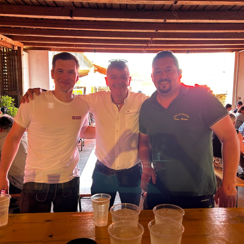 Drei Männer stehen an einem Holztisch mit Getränken davor. Der Mann links trägt ein weißes T-Shirt und grüne Shorts. Der Mann in der Mitte trägt ein beiges Hemd und eine Sonnenbrille auf dem Kopf. Der Mann rechts trägt ein grünes Hemd. Dahinter sitzen mehrere Personen an Tischen.