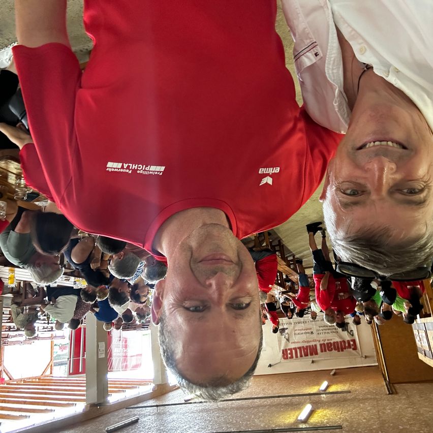 Eine Gruppe von Menschen in roten Shirts, wahrscheinlich bei einem Radsportevent. Ein Mann ist auf dem Kopf stehend, und ein anderer lächelt. Der Raum hat Banner, Treppen und Lichter.
