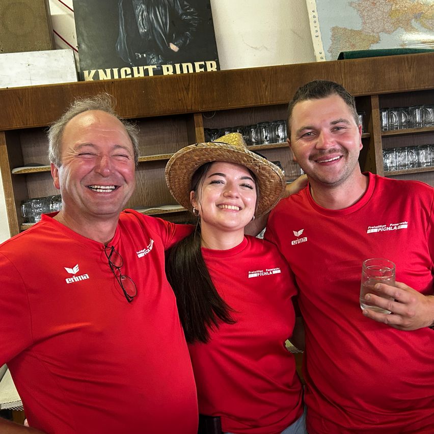 Drei Personen in roten T-Shirts mit Logos lächeln in einem Raum mit Regalen und einer Karte. Die Frau trägt einen Strohhut.