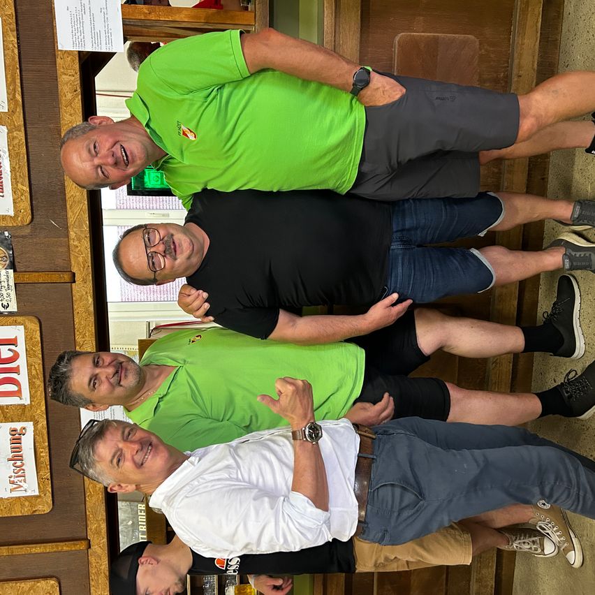 Vier Männer in Freizeitkleidung posieren für ein Foto. Einer trägt ein grünes Poloshirt, zwei tragen Schwarz und einer trägt Weiß. Sie lächeln und stehen eng beieinander.
