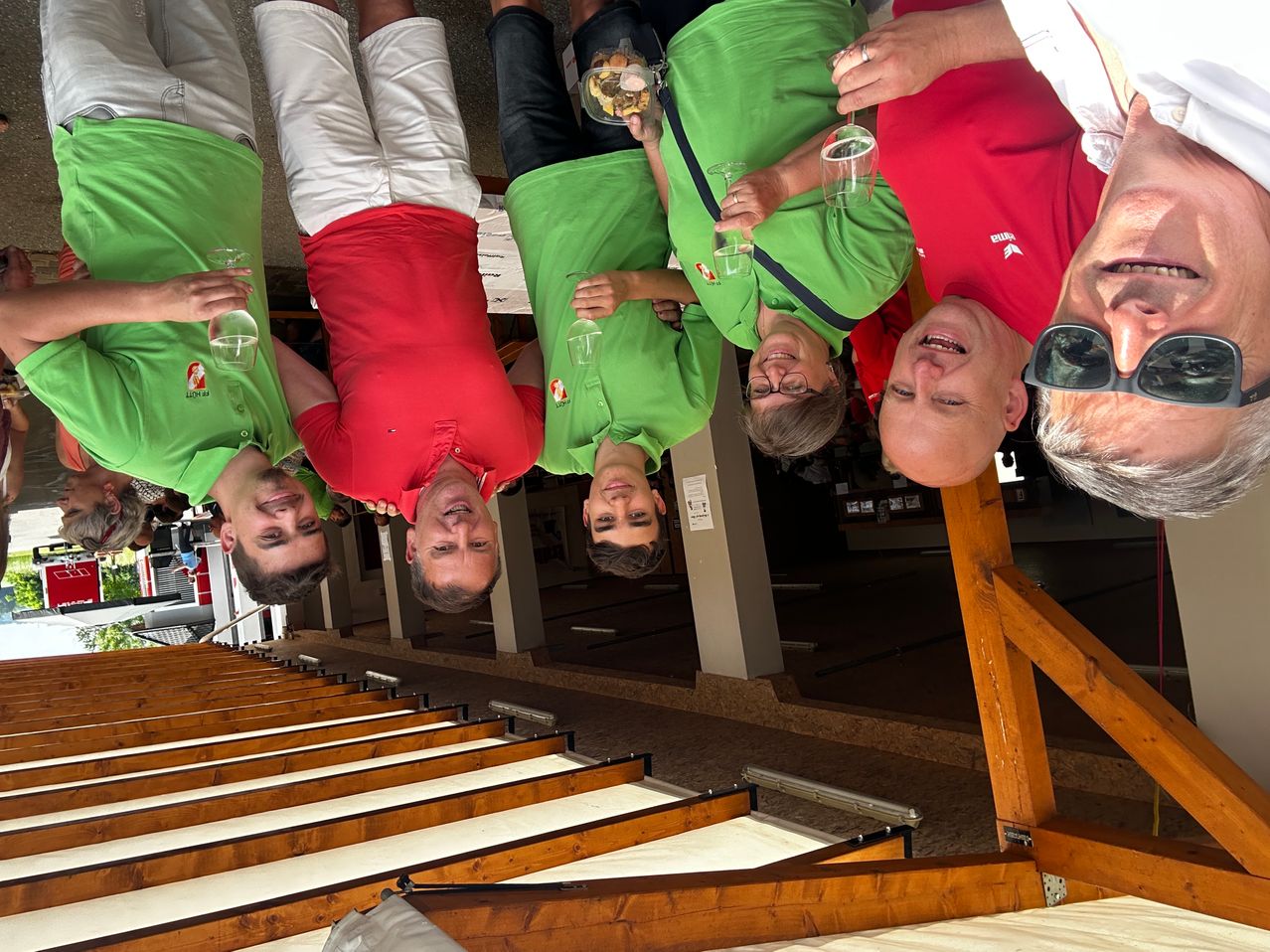 Eine Gruppe von Menschen in grünen und roten Shirts hält Getränke in einem Restaurant. Sie lächeln und scheinen für ein Foto zu posieren. Das Restaurant hat eine Holzdecke.