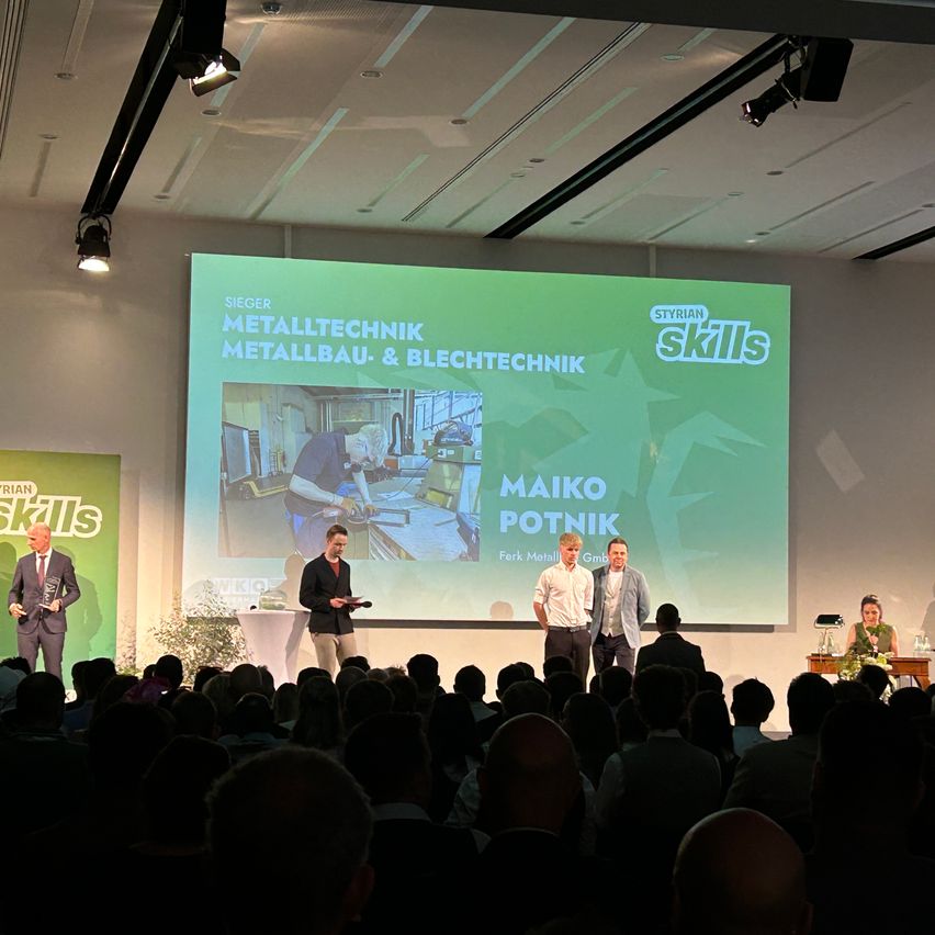 A group of men stand on stage in front of a projector screen showing a presentation about metal technology. The audience is seated in front of them.