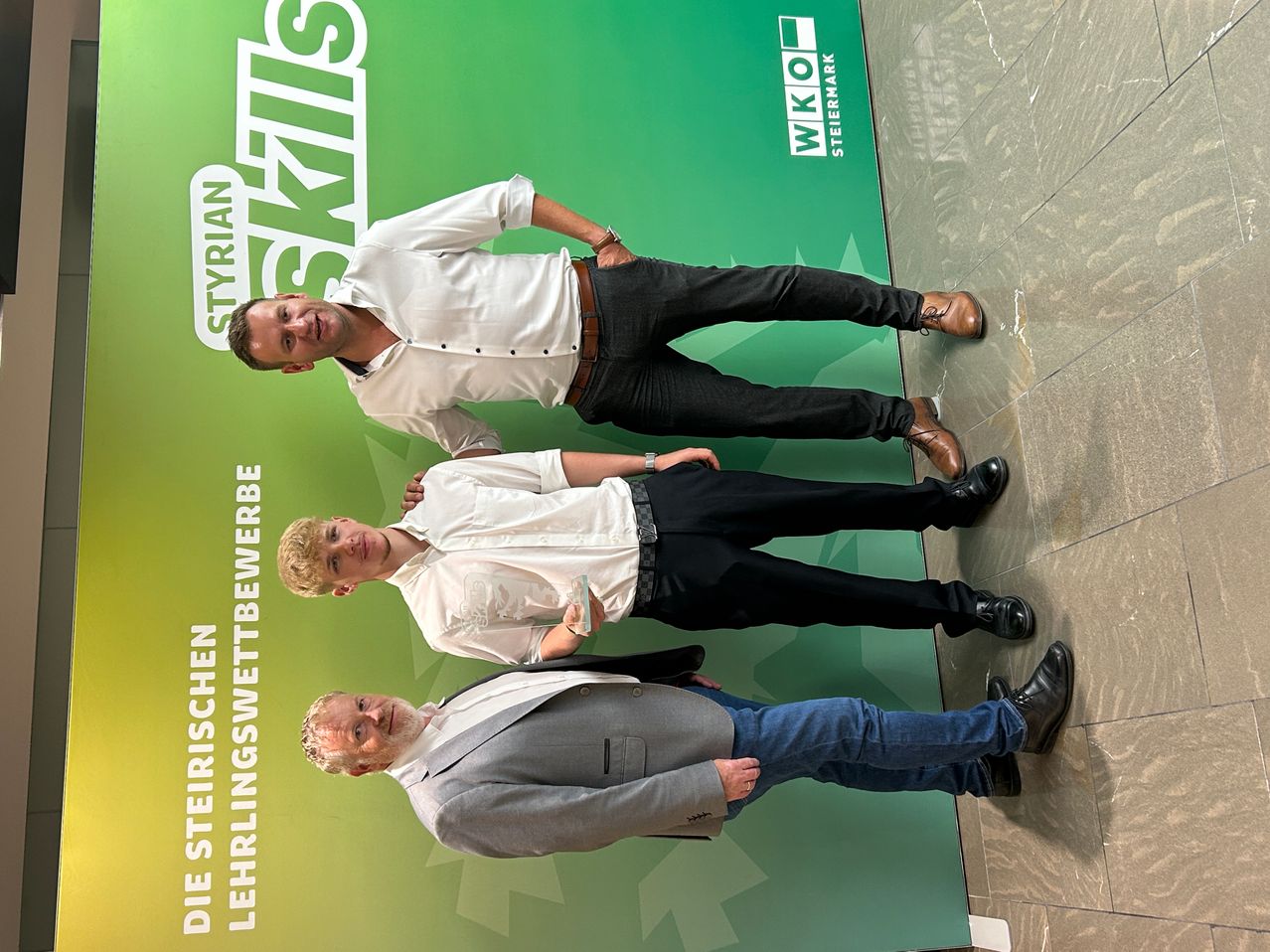 Three men are posing for a photo in front of a green banner. The man in the middle holds an award. The man on the left wears a white shirt, and the man on the right wears a gray suit. The banner has white text on a green background.