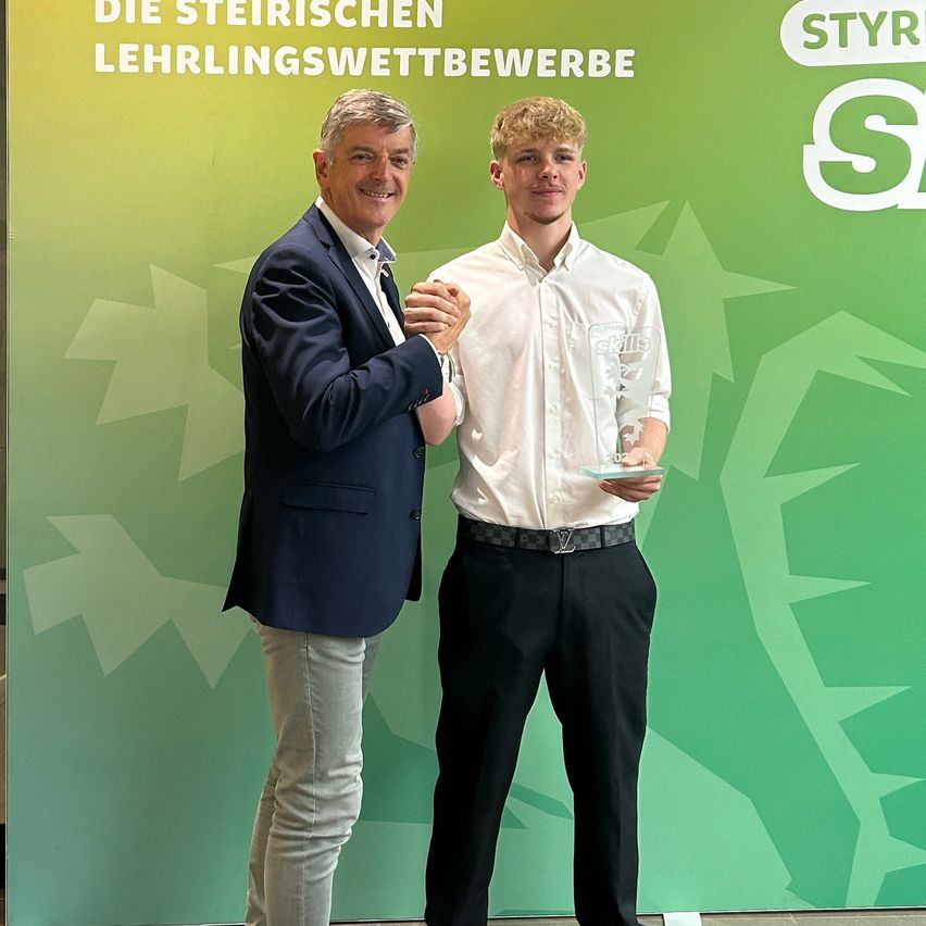 A man in a suit presents an award to a young man in a white shirt holding a trophy. They stand in front of a green banner with white text.