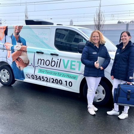 Zwei Frauen stehen vor einem weißen Van mit einer Katze und Ärzten auf der Seite und lächeln. Sie halten beide etwas in den Händen. Der Van hat 'mobilVET' darauf geschrieben.