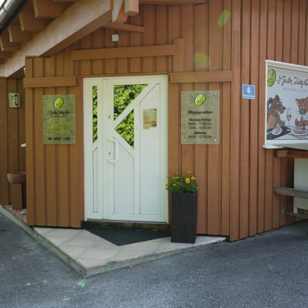 Der Eingang zu einem Holzchalet zeigt Öffnungszeiten an und verfügt über eine weiße Tür, einen Topf mit gelben Blumen und ein Schild für 'Jutta Ziegle Naturfutter'.