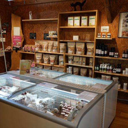 Ein holzfarbener Laden zeigt Regale mit verschiedenen Lebensmitteln und Flaschen. Eine Glasvitrine enthält weitere Lebensmittel. Eine Katzenstatue und ein Schild hängen an der Wand.