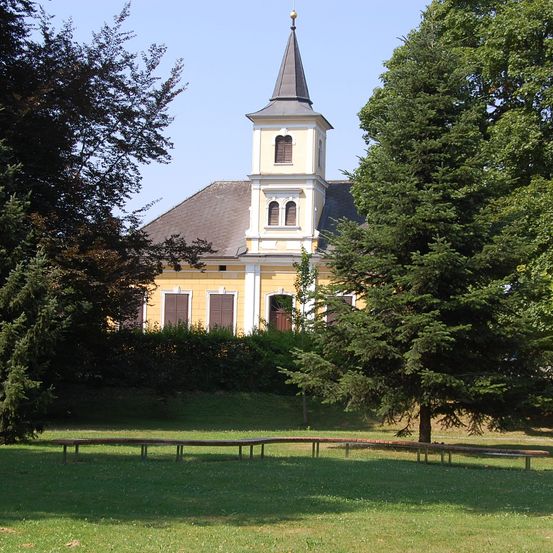 Eine gelbe Kirche mit grauem Dach und einem hohen Turm steht in einem üppigen grünen Park. Bäume und Büsche umgeben das Gebäude, und vor dem Gebäude sind Bänke aufgestellt.