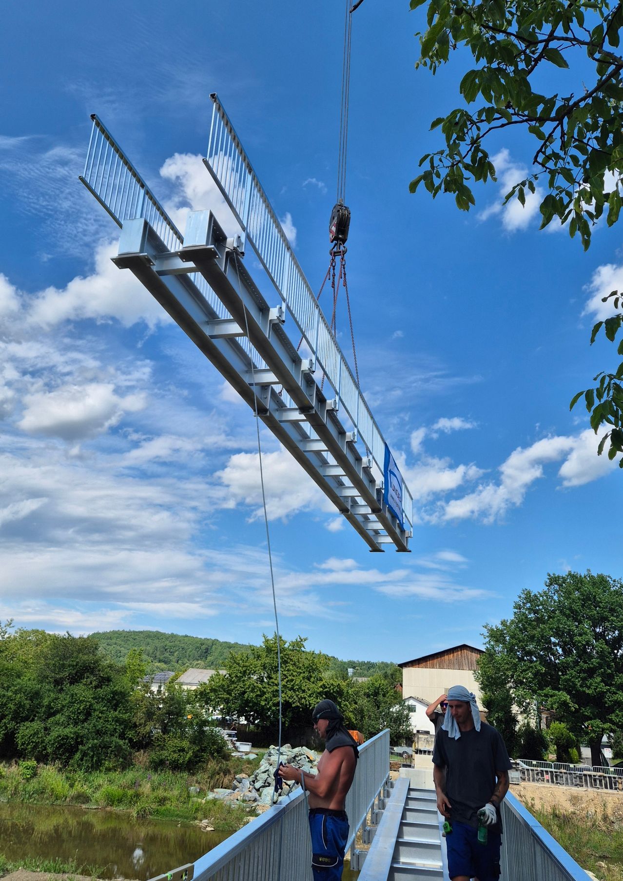Eine Baustelle mit einem metallischen Träger, der von einem Kran gehoben wird. Zwei Personen sind anwesend, eine trägt einen Hut und die andere ein schwarzes T-Shirt. Bäume und ein Haus sind im Hintergrund unter einem klaren blauen Himmel mit verstreuten Wolken zu sehen.