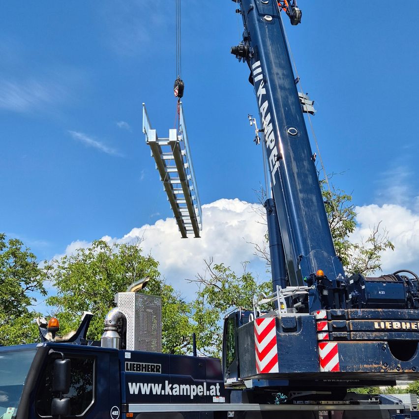 Ein großer Kran hebt eine silberne Leiter unter einem klaren blauen Himmel mit weißen Wolken. Der Kran ist blau mit roten und weißen Streifen und einem Logo. Bäume sind im Hintergrund.