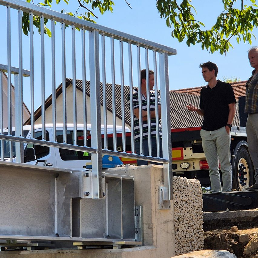 Drei Männer stehen auf einer Baustelle mit einem Metallzaun und einem Lkw im Hintergrund. Ein Mann spricht, während die anderen zuhören.