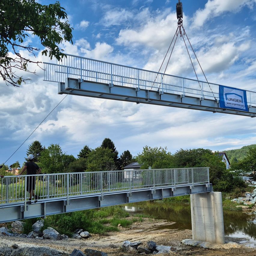 Eine Brücke wird von einem Kran in Position gehoben, wobei eine Person mit einer weißen Helm auf der Brücke steht. Die Brücke hat ein Metallgeländer und ein blaues Banner mit dem Text 'UNGER'. Die Brücke befindet sich über einem Gewässer. Im Hintergrund befinden sich Bäume und ein Haus.