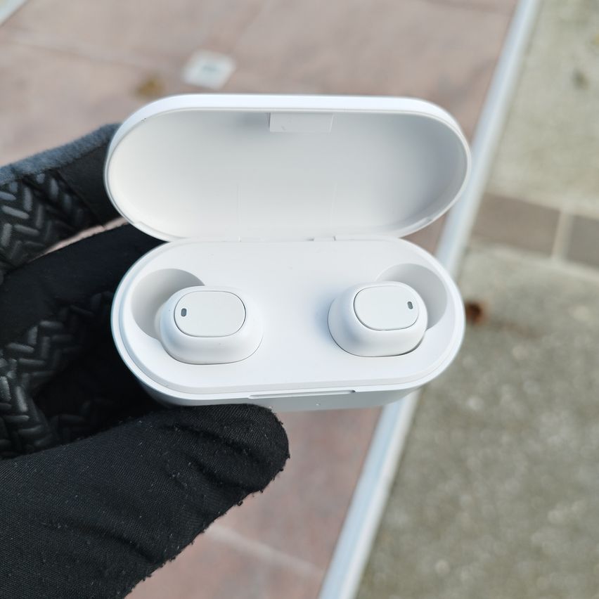 Eine Person mit schwarzen Handschuhen hält ein weißes kabelloses Ohrhörer-Etui mit zwei Ohrhörern im Inneren. Die Ohrhörer haben ein kleines Touchpad an der Seite.