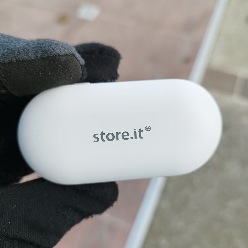 Eine behandschuhte Hand hält ein weißes Objekt mit dem Text 'store.it' in einer fremden Sprache. Das Objekt ist ein kleines, abgerundetes und möglicherweise elektronisches Gerät.