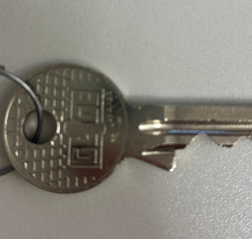 Eine Nahaufnahme eines silbernen Schlüssels mit Schlüsselloch, der ein geometrisches Muster auf dem Kopf und eine gebogene Spitze aufweist. Der Schlüssel ist an einem Schlüsselbund befestigt.