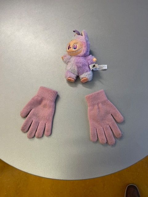 Ein kleines lila Plüschtier mit großen Augen liegt auf einem Tisch. Zwei rosa Handschuhe daneben. Die Handschuhe sind zur Hälfte gefaltet. Der Tisch hat eine Metalloberfläche.