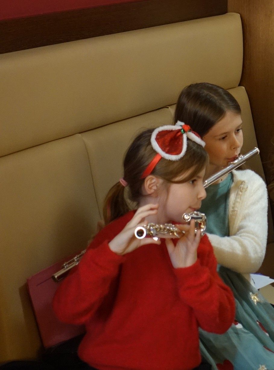 Zwei junge Mädchen sitzen auf einer Bank und spielen Flöten. Das Mädchen in Rot hat eine festliche Haarschleife. Sie befinden sich wahrscheinlich in einem Klassenzimmer oder Musikstudio.