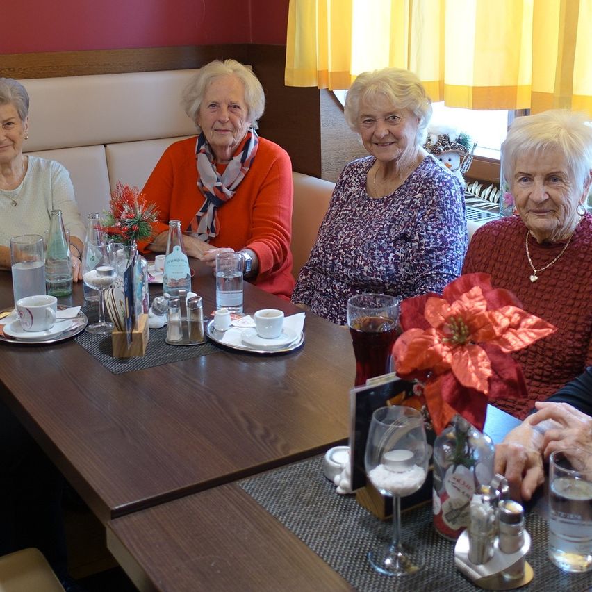 Vier ältere Frauen sitzen lächelnd an einem Tisch. Der Tisch hat Tassen, Untertassen, eine Flasche und eine Blume. Sie scheinen sich in einem Restaurant zu befinden.