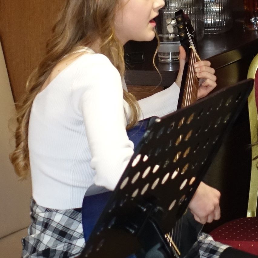 Ein junges Mädchen mit langen blonden Haaren spielt Gitarre. Sie singt und hält die Gitarre. Sie sitzt vor einem Notenständer. Dahinter ist eine verschwommene Ansicht eines hölzernen Schranks.