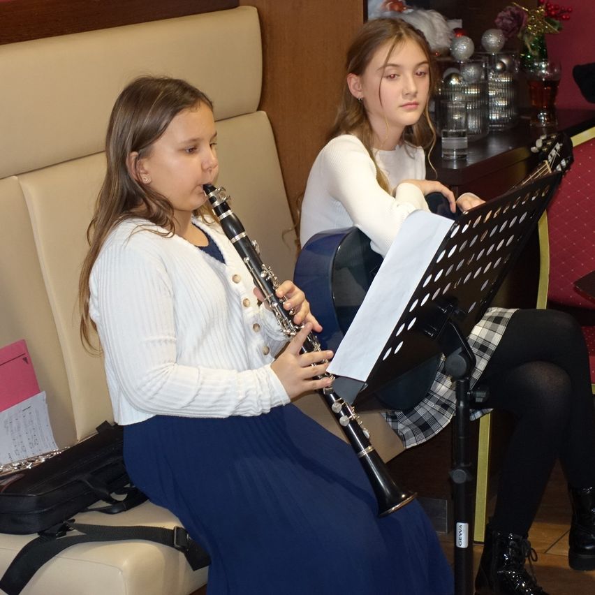 Zwei junge Mädchen sitzen, eine spielt Klarinette und die andere Gitarre, mit Notenblättern vor ihnen. Sie sind in Winterkleidung gekleidet und scheinen sich in einem Restaurant zu befinden.