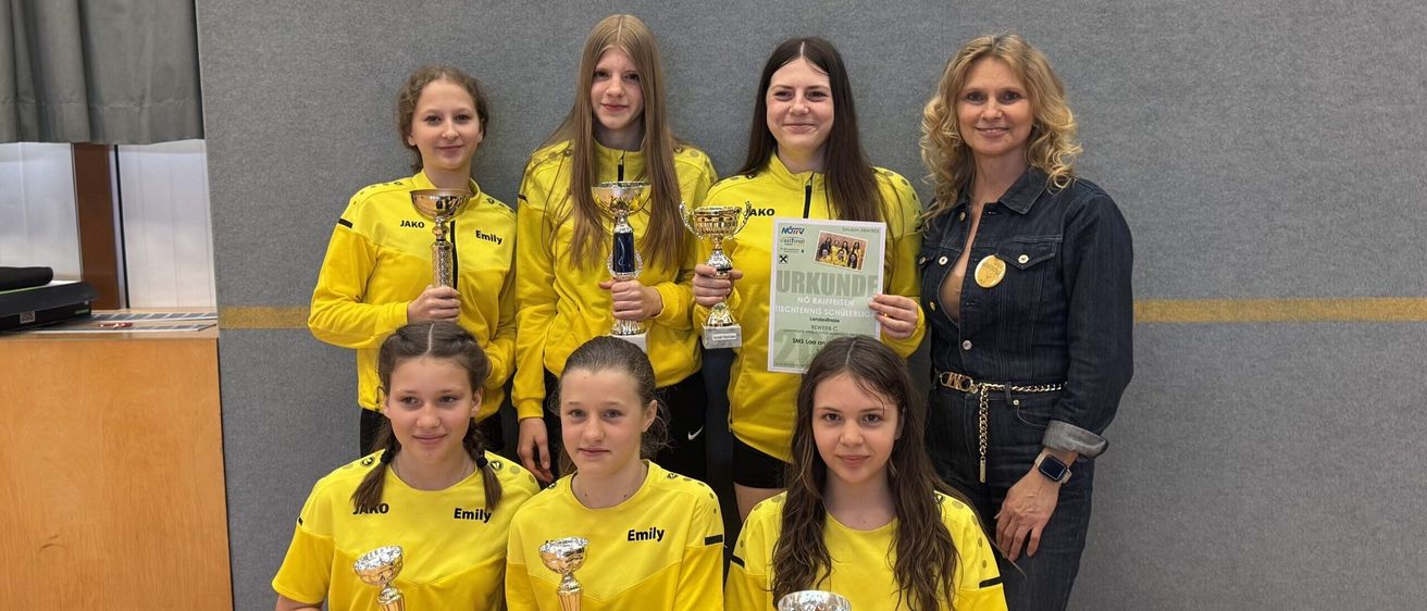 Eine Gruppe von Mädchen in gelben Trikots mit dem Wort Emily und halten Pokale. Sie stehen vor einer grauen Wand mit einer Frau in einer Jeansjacke und einer Uhr.