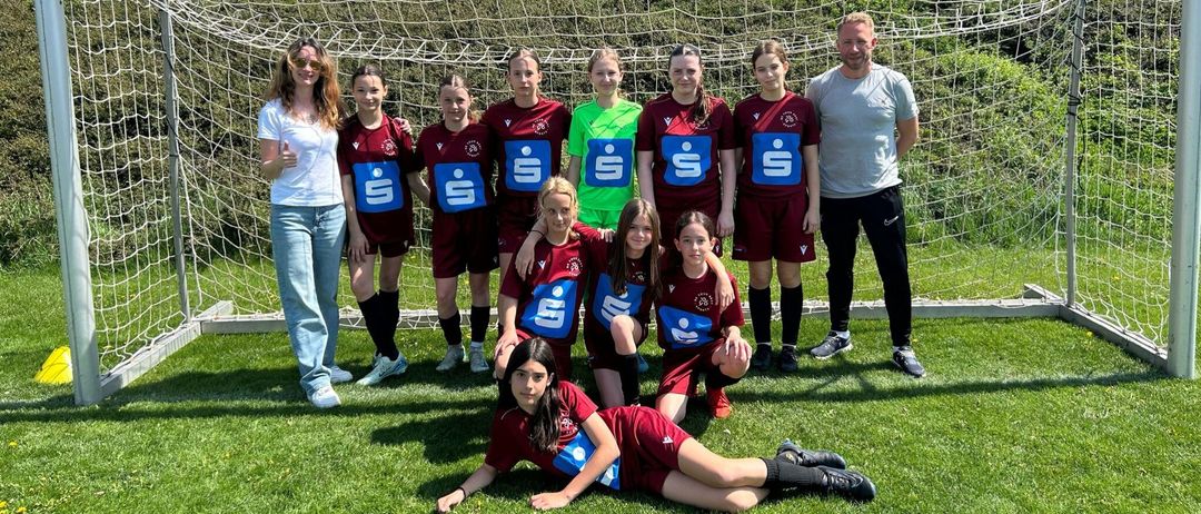 Eine Frauen-Fußballmannschaft posiert für ein Foto vor einem Tor, wobei einige Spielerinnen auf dem Gras knien.