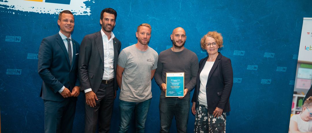 Fünf Personen stehen nebeneinander. Der Mann in der Mitte hält eine Plakette, mit einer Frau zu seiner rechten und drei Männern zu seiner linken Seite. Sie sind alle schick gekleidet und stehen vor einer blauen Wand.