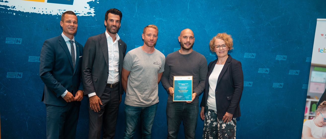 Fünf Personen stehen nebeneinander. Der Mann in der Mitte hält eine Plakette, mit einer Frau zu seiner rechten und drei Männern zu seiner linken Seite. Sie sind alle schick gekleidet und stehen vor einer blauen Wand.
