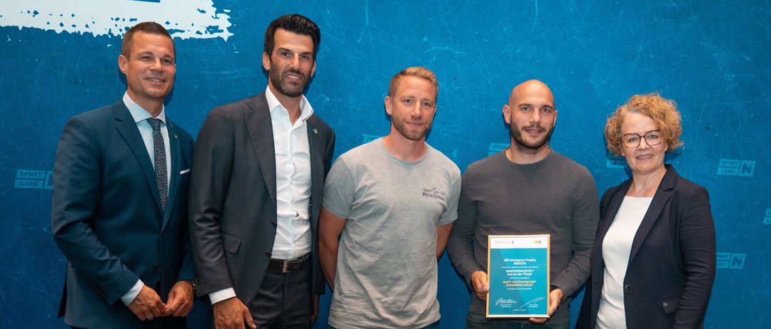 Fünf Männer stehen vor einer blauen Wand. Ein Mann hält ein blaues Zertifikat. Ein anderer Mann trägt ein graues T-Shirt.