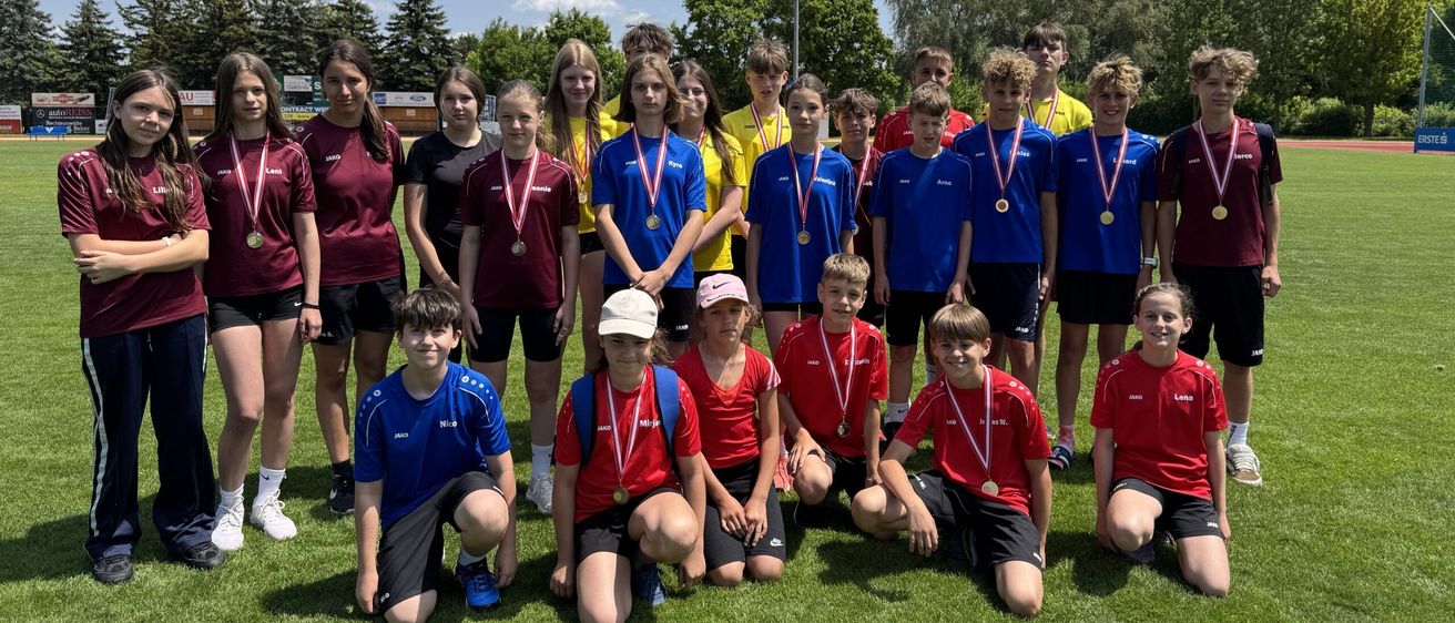 Eine Gruppe junger Athleten in Teamuniformen posiert für ein Foto auf einem Feld, mit Medaillen um den Hals, mit Bäumen und einer Straßenlaterne im Hintergrund.