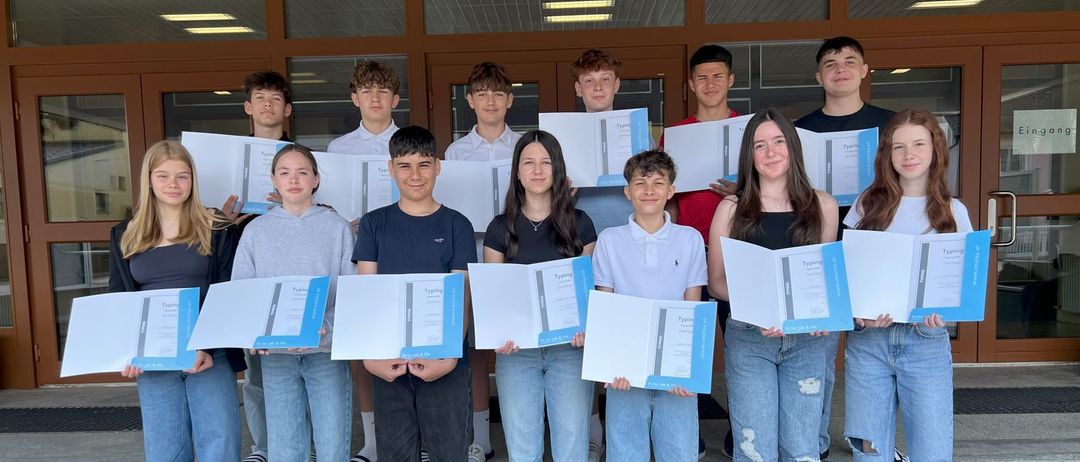 Eine Gruppe junger Menschen posiert für ein Foto auf einer Treppe, hält Zertifikate in der Hand und lächelt. Der Raum hat Holztüren und Glasfenster.