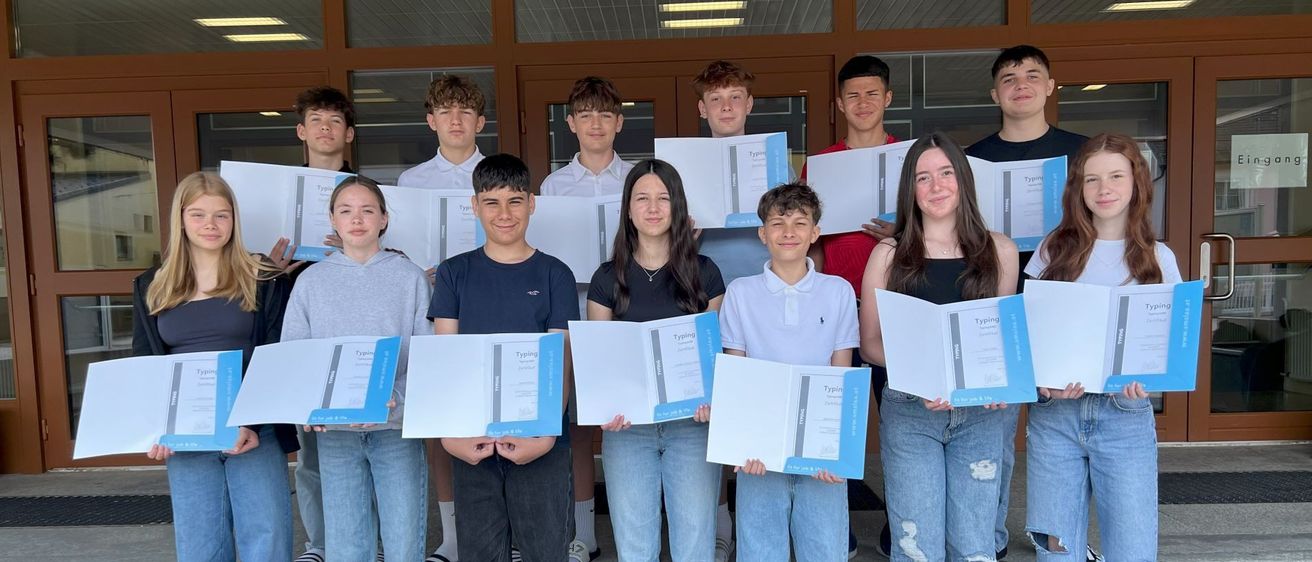 Eine Gruppe junger Menschen posiert für ein Foto auf einer Treppe, hält Zertifikate in der Hand und lächelt. Der Raum hat Holztüren und Glasfenster.