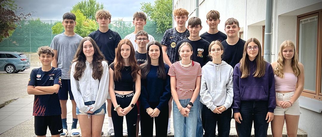 Eine Gruppe junger Menschen, Jungen und Mädchen, posiert für ein Foto auf einer Treppe. Sie tragen Freizeitkleidung. Bäume und ein Gebäude sind im Hintergrund.