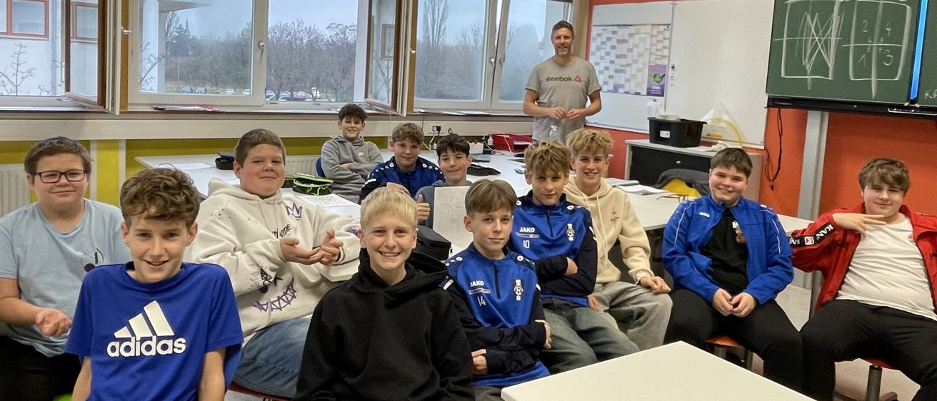 Mehrere Jungen sitzen in einem Klassenzimmer und lächeln für ein Foto. Ein Mann steht am Fenster, und der Raum hat Schreibtische und eine Tafel.