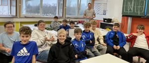 Mehrere Jungen sitzen in einem Klassenzimmer und lächeln für ein Foto. Ein Mann steht am Fenster, und der Raum hat Schreibtische und eine Tafel.