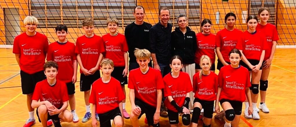Eine Gruppe junger Athleten in roten Trikots posiert mit zwei Männern und zwei Frauen für ein Foto in einer Turnhalle. Die Turnhalle hat einen Basketballkorb und ein Netz.