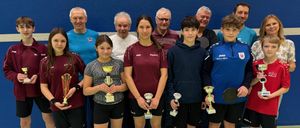 Eine Gruppe junger Athleten, sowohl Jungen als auch Mädchen, stehen zusammen und halten Trophäen. Sie tragen Sportuniformen und lächeln für ein Foto. Hinter ihnen stehen ältere Männer, wahrscheinlich ihre Trainer, die ebenfalls lächeln. Sie stehen vor einer blauen Wand.
