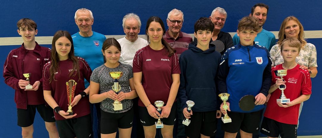 Eine Gruppe junger Athleten, sowohl Jungen als auch Mädchen, stehen zusammen und halten Trophäen. Sie tragen Sportuniformen und lächeln für ein Foto. Hinter ihnen stehen ältere Männer, wahrscheinlich ihre Trainer, die ebenfalls lächeln. Sie stehen vor einer blauen Wand.