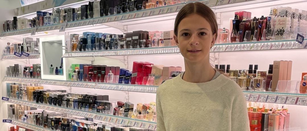 Eine junge Frau steht in einem Kosmetikgeschäft, einen Einkaufswagen haltend, umgeben von Regalen voller verschiedener Düfte und Schönheitsprodukte.