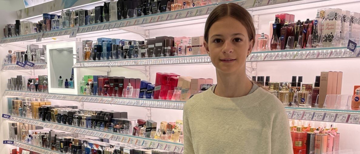 Eine junge Frau steht in einem Kosmetikgeschäft, einen Einkaufswagen haltend, umgeben von Regalen voller verschiedener Düfte und Schönheitsprodukte.