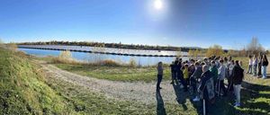 Eine Gruppe von Schülern steht am Ufer eines Sees mit einem Solarpark dahinter und hört einer Frau zu, die an einem blauen Himmel spricht.