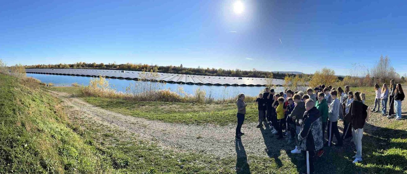 Eine Gruppe von Schülern steht am Ufer eines Sees mit einem Solarpark dahinter und hört einer Frau zu, die an einem blauen Himmel spricht.