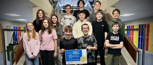 Eine Gruppe junger Schüler posiert auf einer Treppe für ein Foto, einer hält ein Zertifikat und lächelt. Hinter ihnen trägt ein weiterer Schüler eine 'Jack & Jones' Kapuzenjacke.