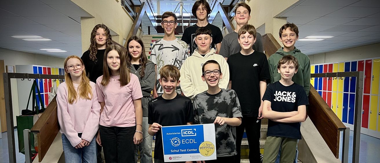 Eine Gruppe junger Schüler posiert auf einer Treppe für ein Foto, einer hält ein Zertifikat und lächelt. Hinter ihnen trägt ein weiterer Schüler eine 'Jack & Jones' Kapuzenjacke.