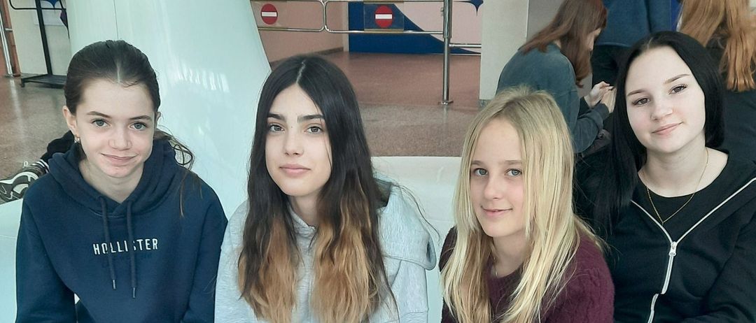 Vier junge Frauen posieren für ein Foto auf einer Bank. Die Frau auf der linken Seite trägt eine blaue Kapuzenjacke mit weißen Buchstaben. Die Frau neben ihr trägt eine graue Kapuzenjacke. Die dritte Frau trägt ein kastanienbraunes Oberteil. Die vierte Frau trägt eine schwarze Jacke.