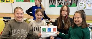 Vier Mädchen sitzen in einem Klassenzimmer auf Stühlen und lächeln. Eines hält ein Papier, wahrscheinlich ein Zertifikat, während die anderen zuschauen. Bunte Kunstwerke schmücken die Wände.