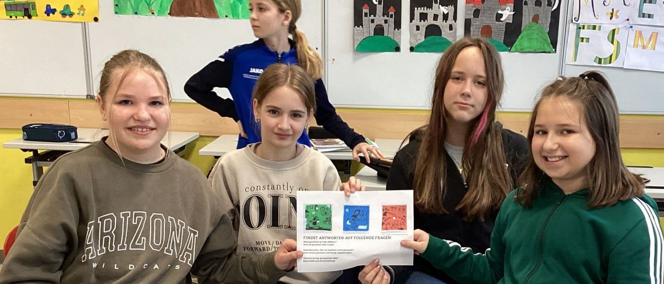 Vier Mädchen sitzen in einem Klassenzimmer auf Stühlen und lächeln. Eines hält ein Papier, wahrscheinlich ein Zertifikat, während die anderen zuschauen. Bunte Kunstwerke schmücken die Wände.