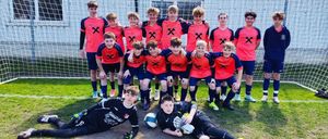 Eine Fußballmannschaft posiert für ein Gruppenfoto vor einem Netz, Spieler in orangefarbenen und blauen Trikots. Einige stehen, andere liegen auf dem Boden, alle lächeln.