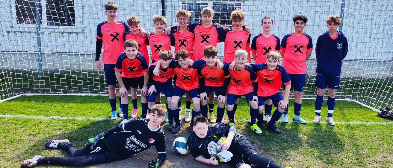 Eine Fußballmannschaft posiert für ein Gruppenfoto vor einem Netz, Spieler in orangefarbenen und blauen Trikots. Einige stehen, andere liegen auf dem Boden, alle lächeln.